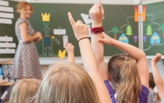 Im verschwommenen Hintergrund steht eine Lehrerin vor einer Tafel. Im fokussiertem Vordergrund melden sich Kinder