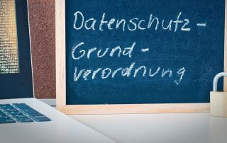 DSGVO Datenschutzerklärung in Kreide auf eine Tafel geschrieben, welche neben einem Laptop steht. Davor steht ein kleines Aufhängeschloss