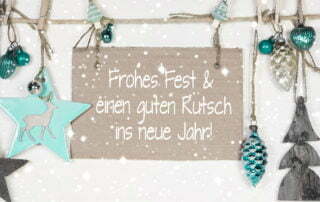 Weihnachtliche Dekoration aus verschiedenen Anhängern, die an einer Schnur befestigt sind. Im Mittelpunkt hängt ein braunes Schild mit der Aufschrift Frohes Fest & einen guten Rutsch ins neue Jahr